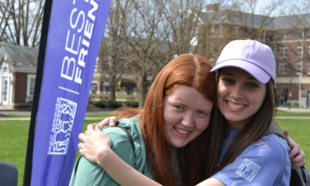 Disabled friendship program: Standing beside a blue "Best Buddies" banner, a dark-haired young woman in a white baseball cap and blue shirt and a red-headed young woman in a green sweatshirt hoodie hug each other.