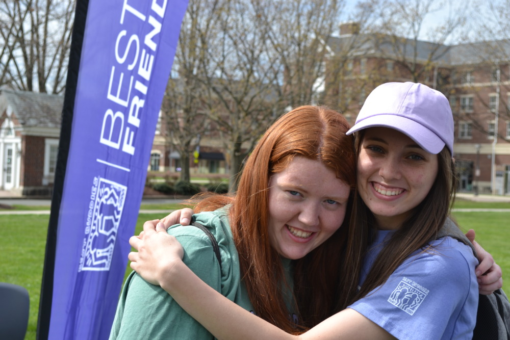 Disabled friendship program: Standing beside a blue "Best Buddies" banner, a dark-haired young woman in a white baseball cap and blue shirt and a red-headed young woman in a green sweatshirt hoodie hug each other.