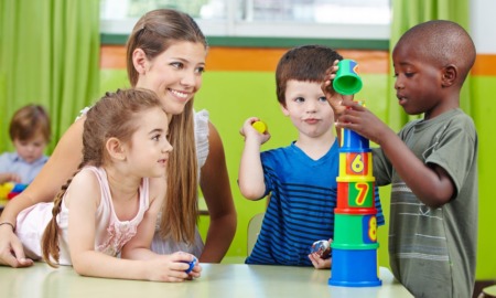 child care research grants: young, female child care professional with three young kids building blocks