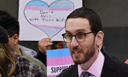 Lawmakers want legal refuge for trans youth: dark-haired man in glasses and suit speaking with trans rights protest sign in background
