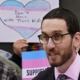 Lawmakers want legal refuge for trans youth: dark-haired man in glasses and suit speaking with trans rights protest sign in background