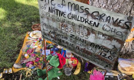Native American Boarding School Deaths: Handprinted sign saying "Site mass graves native children,"sits amidst flowers
