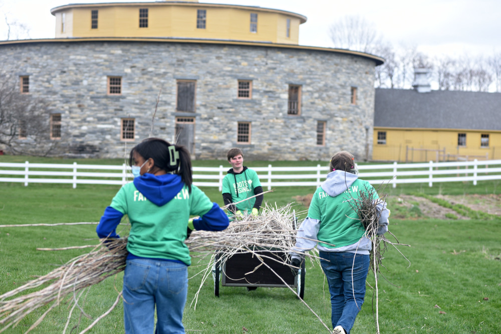 Teen farm program: Three teens wearing light green swatshirts and jeans woking in white fenced farm field with 2 story stone and yellow barn in the background