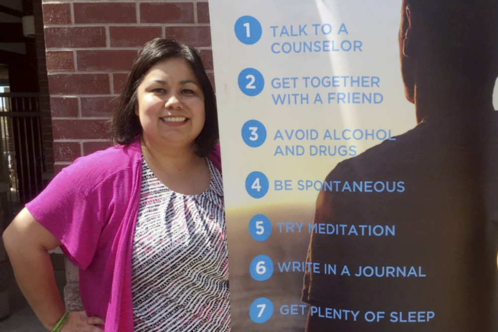 School Counselors and school shootings: Woman with short dark hair weaing pink top over print dress stands next to list of advice for students on a large banner