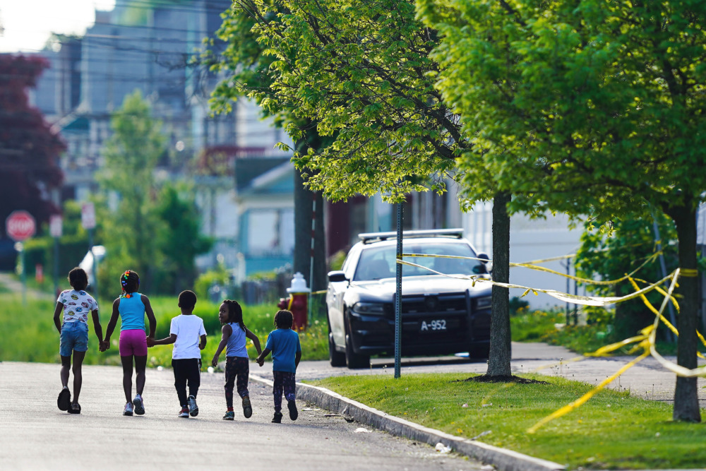 School Counselors and school shootings: Five young children holding hands walk down street passing by the yellow crime tape wrapped around trees on a grass lawn with parked police car.
