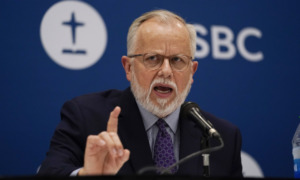 Southern Baptist Offender Man with grey hair and beard wearing dark suit, gestures with right hand while speaking into microphone at podium standing in front of blue SBC banner