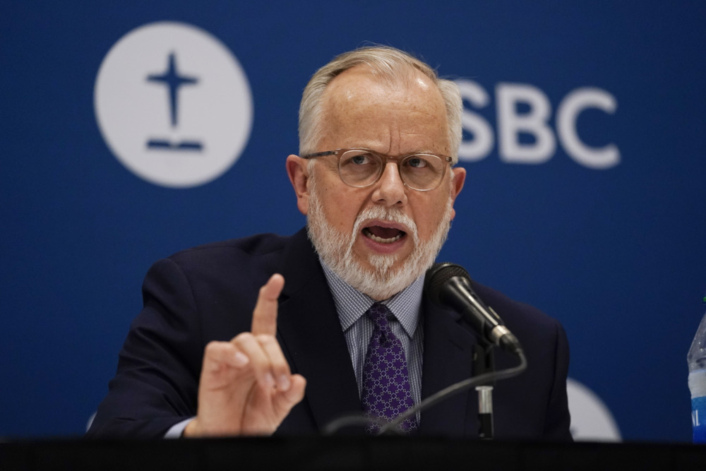 Southern Baptist Offender Man with grey hair and beard wearing dark suit, gestures with right hand while speaking into microphone at podium standing in front of blue SBC banner