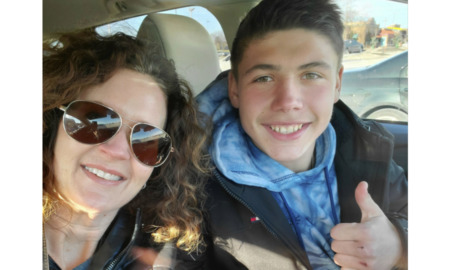 Ukraine war and adoption: Dark-haired woman wearing sunglasses and teen bot with dark hair wearing a dark winter jacker and light blue scark giving thumbs up sign sit in front seats of car smiling into camera.