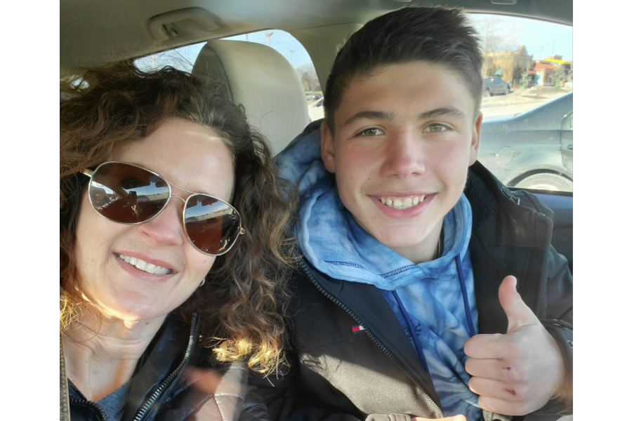 Ukraine war and adoption: Dark-haired woman wearing sunglasses and teen bot with dark hair wearing a dark winter jacker and light blue scark giving thumbs up sign sit in front seats of car smiling into camera.
