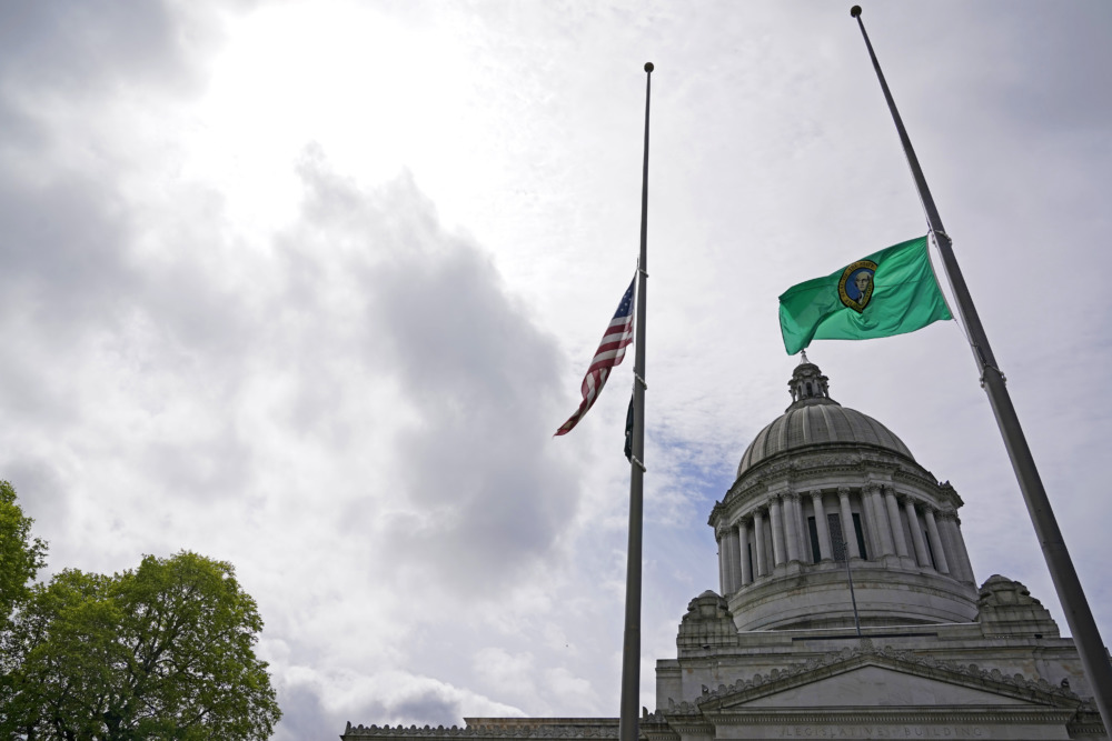Gun control across the nation: AMerican flag and a green state flag at half mast against a cloudy sky in front of a domed tan stone building.