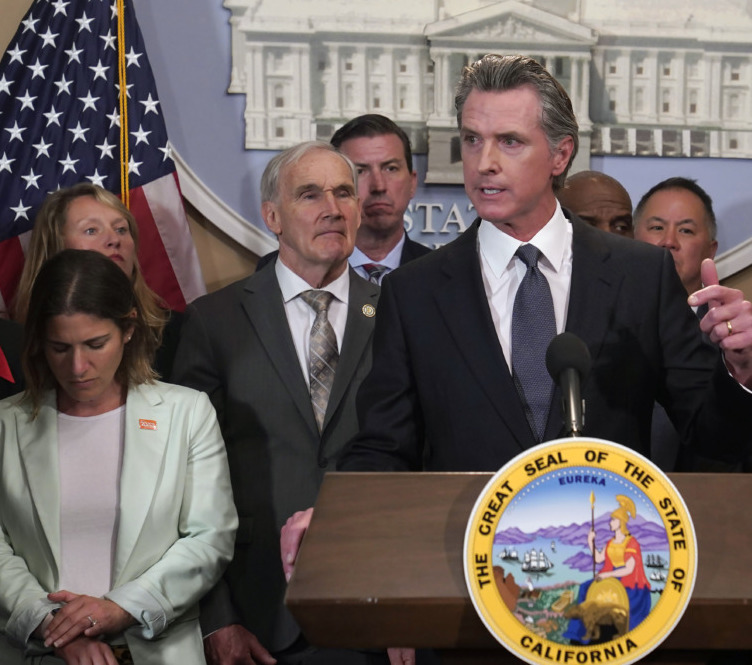 Gun control across the nation: Gray-haired man in navy suit stands behind podium with California gold and blue state seal speaking into microphone with several men in suits standing behind him.