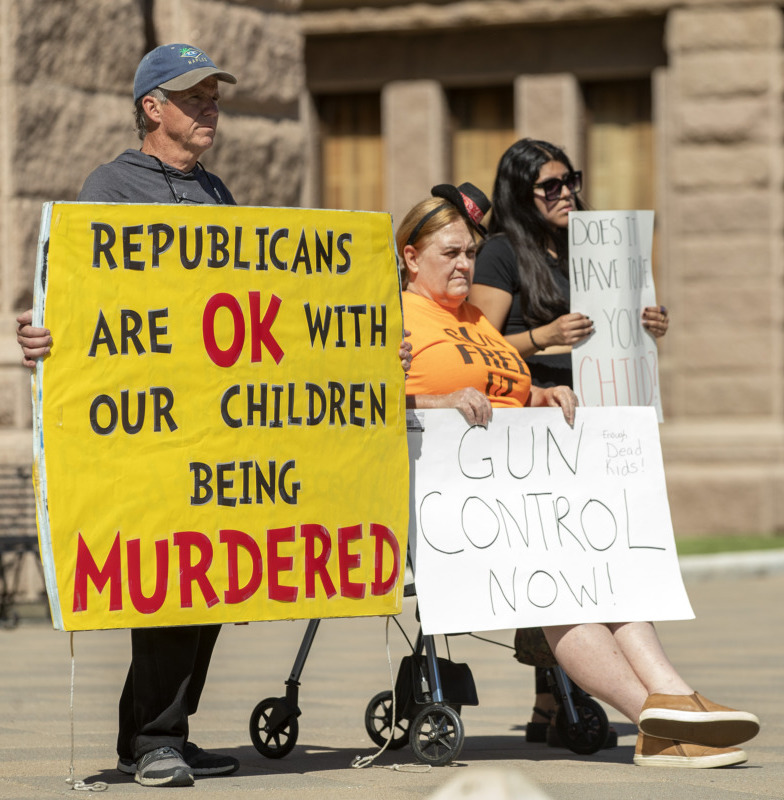 Gun control across the nation: Four adults march in front of a tan stone building carriyng signs, "Republicans are OK with our children being murdered" and "Gun control now!"