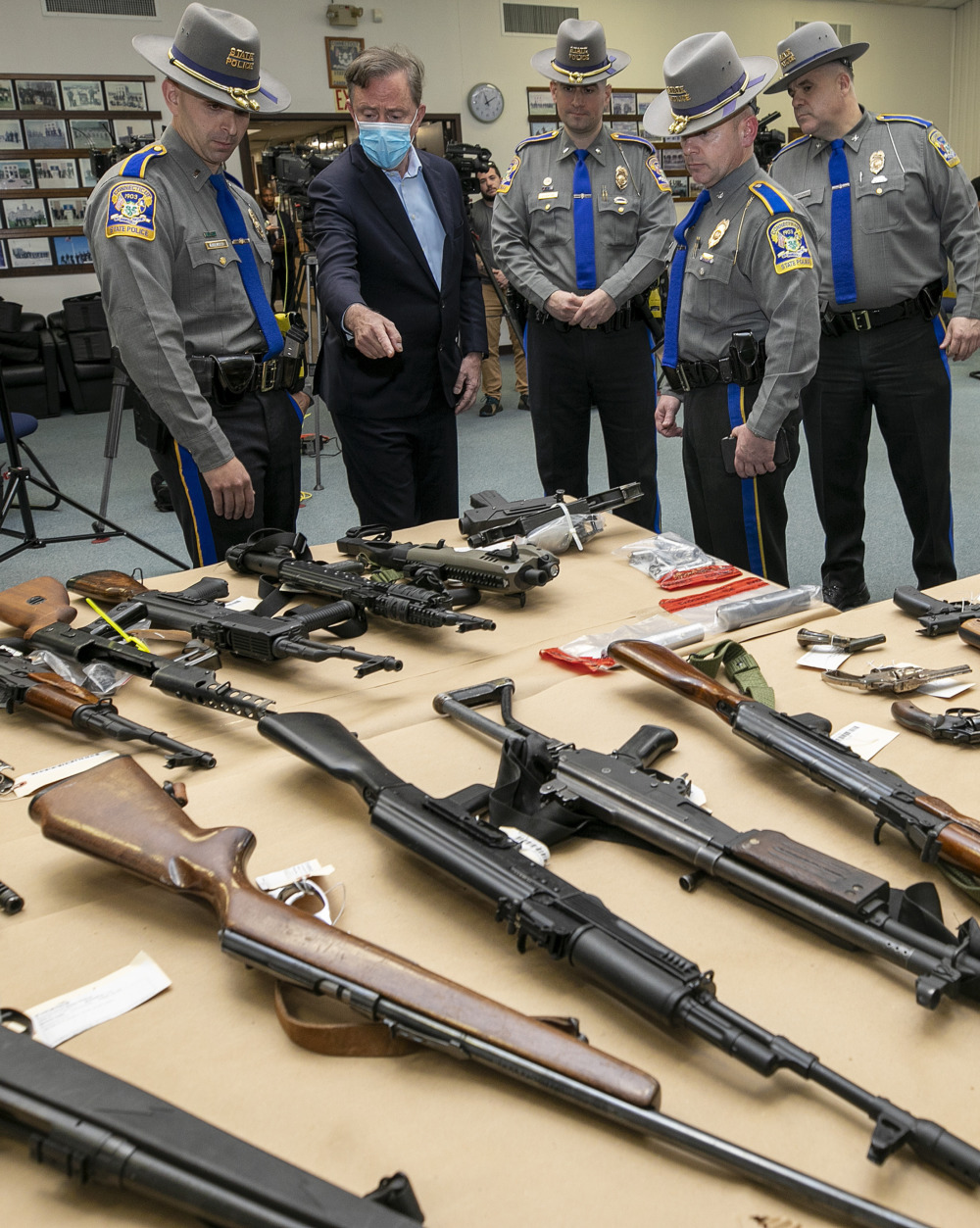 Gun control across the nation: Several men in law enfrement uniforms and one man in a dark suit stand looking down at two tables where numerous guns are laid out.