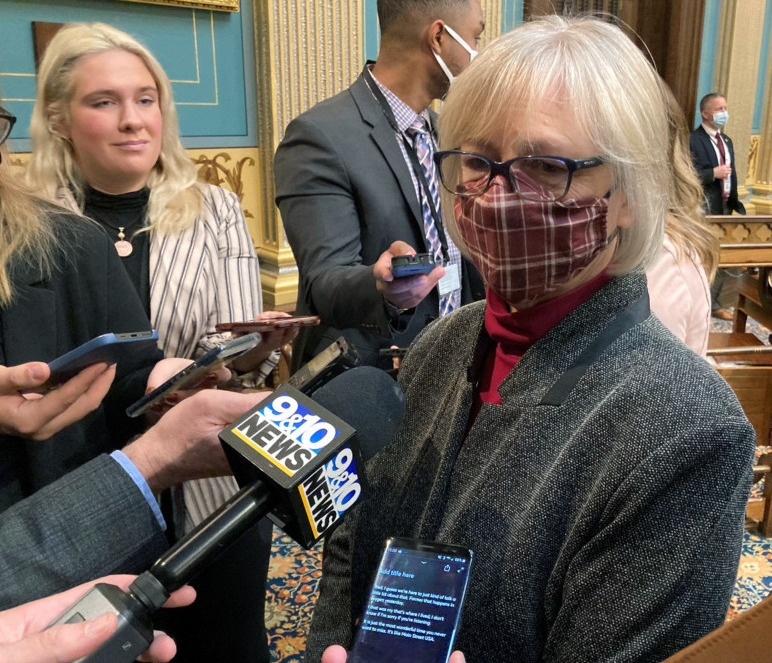 Gun control across the nation: White-haired woman with glasses in gray suit with red top stands speaking nto microphones held by reporters off-camera.