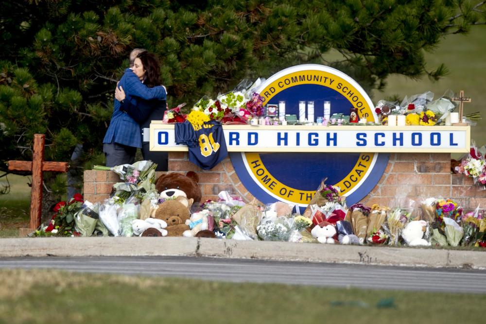 school shooting: Oxford high school sign covered in flowers and memorial items while two people hug