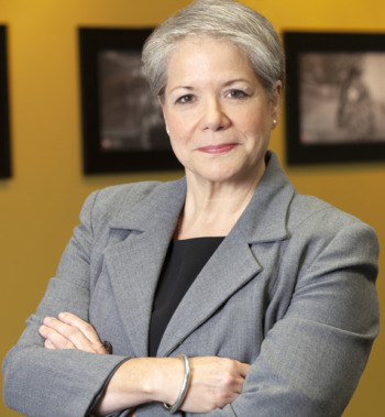 Online adoption galleries: A woman with gray hair pulled back earing grey jacket and block top stands with arms crossed in front of a yellow wall