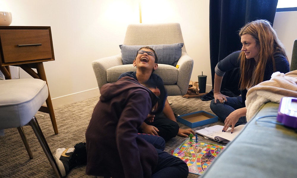 young caregivers offer crucial help: young boy laughs while playing games with another boy and woman in hotel room