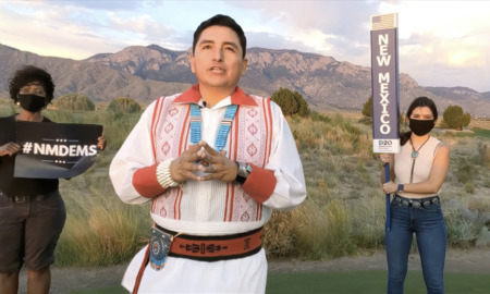 Native indigenous language: Man with black hair pulled back wearing native clothing with re detailing on white shirt stands in desert landscape i front of two adults holding signs