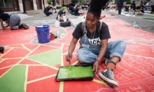 NYC community grants; arts and culture project grants: young black woman in overall painting on ground with lots of others painting in background