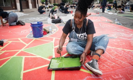 community arts and culture project grants: young black woman in overall painting on ground with lots of others painting in background