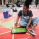 community arts and culture project grants: young black woman in overall painting on ground with lots of others painting in background