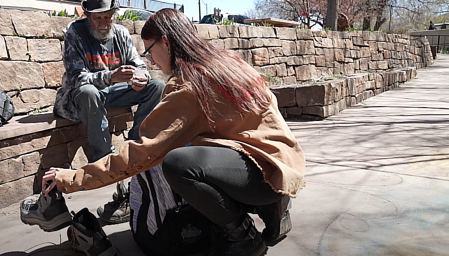 Homeless youth in New Mexico: young,woman with long dark hair wearing black pants and tan jacket crouches on sidewalk showing pairs of used sneakers to older homeless man with gray beard wearing a hat, jeans and plaid shirt sitting on stone bench.