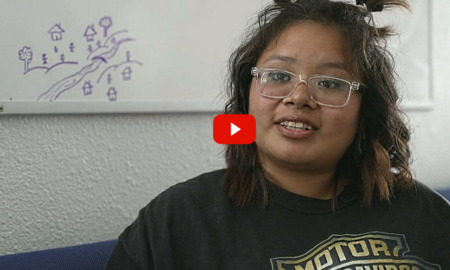 New Mexico Homeless youth: Young woman with dark hair and clear-framed glasses wearing black sweatshirt with Harlet-Davidson graphic in gold sits in couch speaking into camera