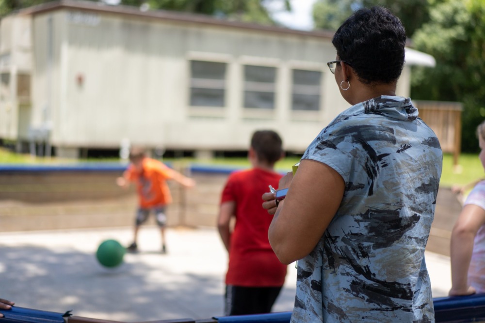 YMCA summer program: Woman stands to far right with back to camera as tatching Two children face each as one prepares to kick approaching kickball.