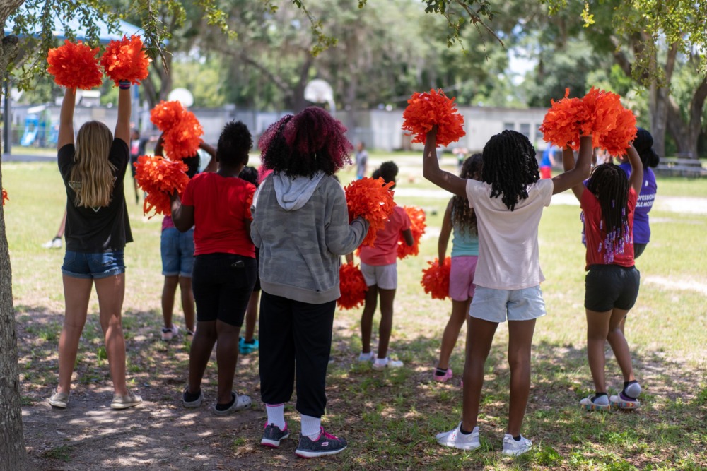 YMCA summer program: Several children in t-shirts and shorts holding orange/red pompoms in the air stand facing a grassy sports field with backs to camera