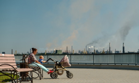 Childhood asthma: Woman sits on a park bench, while baby is in stroller, with industrial smokestacks in the background.