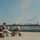 Childhood asthma: Woman sits on a park bench, while baby is in stroller, with industrial smokestacks in the background.