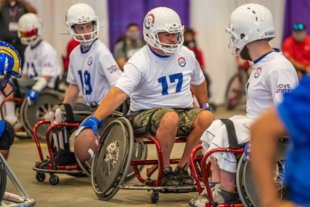 Wheelchair football: Several players from one team in wheelchairs wearing white and blue team shirts and helmets prepare to hike a football