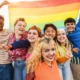 LGBTQ+ community: Young diverse people having fun holding LGBT rainbow flag outdoor - Focus on center blond girl