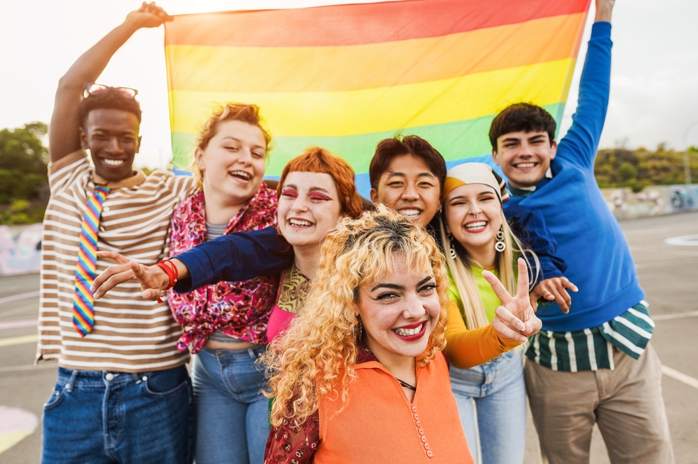 LGBTQ+ community: Young diverse people having fun holding LGBT rainbow flag outdoor - Focus on center blond girl