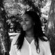 College pandemic fallout: TYoung black woman with long black hair wearing a white blouse stands outside surrounded by large trees.