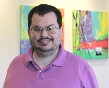 Hotline autism training: Man with dark brown hair, beard and metal frame glasses wearing a lavender polo shirt stands in front of two colorful abstract painting