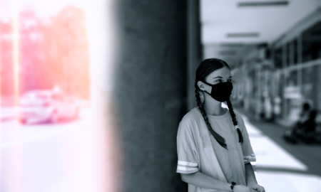 Hotline autism training: Teen girl with long dark braids, black face mask and oversized gray t-shirt stands alone leaning against a cement pillar under an overhang on sidewalk of bsy strret