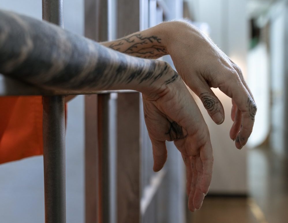 Girl vs. boy juvenile incarceration: A person's tattooed forearms and hands dangle between cell bars.