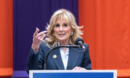 Jill Biden education: Middle-aged blonde woman in navy suit stands speaking into microphone at podium in front of red and blue curtain