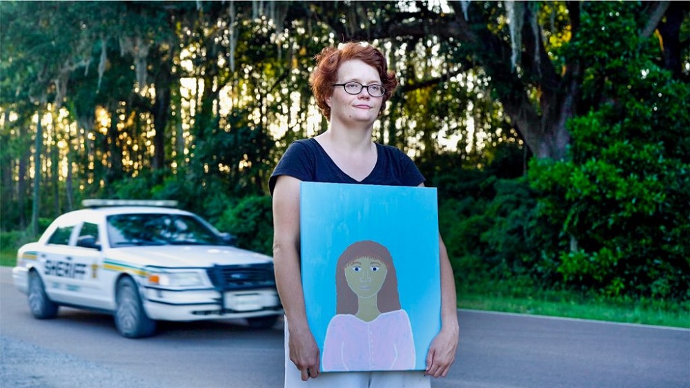 Being Michelle documentary: woman with short hair and glasses stands in road holding a painting of woman