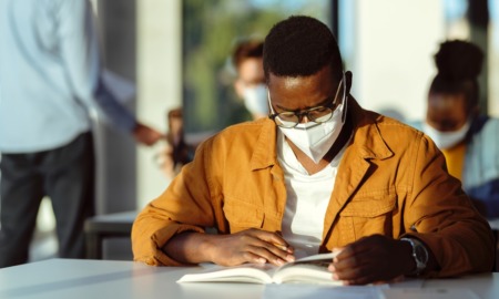 COVID grads face college: Black student with glasses and facemask looks down at schoolwork in a classroom