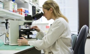 Women in STEM grants, STEM education grants: young blonde woman in lab coat looking into microscope