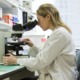 STEM education grants: young blonde woman in lab coat looking into microscope