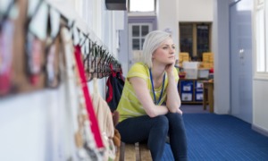 teacher mental health support: young woman teacher sits on bench in school hallway looking out window in thought