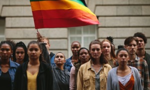 LGBTQ+ community youth: group of stoic youth standing with one holding a rainbow flag