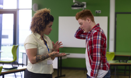 COVID after effects: Black woman with tattoo on right forearm wearing white blouse and key lanyard ID gesturing with hands stands facing white teen boy wearing red plaid shirt