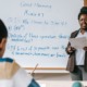 Teacher shortage: Black woman stands in front of white board with math problems dacing desks of elementary students.