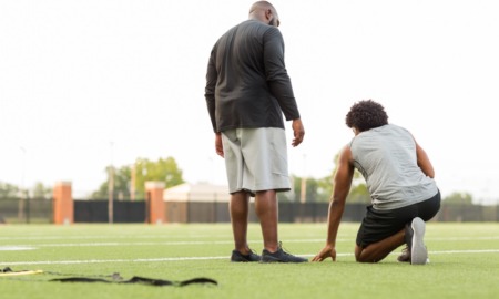 physical education: bald black man stands next to young black student kneeling on football field