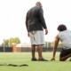 physical education: bald black man stands next to young black student kneeling on football field