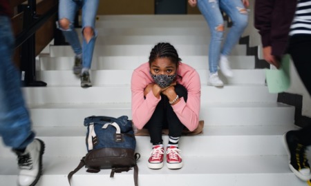 2021 national school climate survey: young black student with facemask sitting on steps sadly while other students walk by